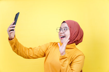 A cheerful asian woman in a hijab and glasses takes a selfie while waving, smiling, and looking at...