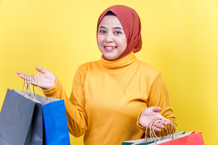 A happy asian woman in a hijab holds colorful shopping bags, smiling confidently against a bright yellow background.の写真素材