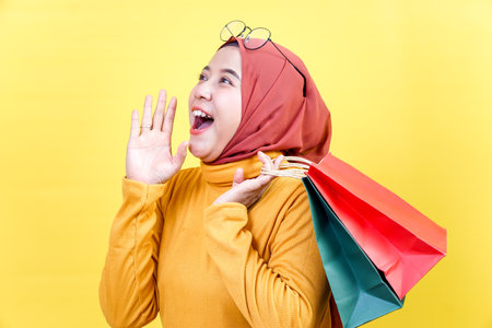 A cheerful asian woman in a hijab excitedly announces a shopping deal while holding colorful bags.