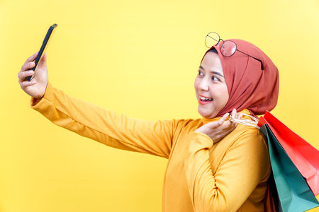 A joyful asian woman in a hijab takes a selfie while holding shopping bags, celebrating a fun...