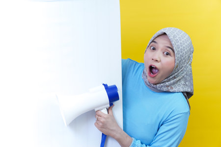 A shocked asian woman in a hijab playfully peeks from behind a white wall, holding a megaphone against yellow.の写真素材