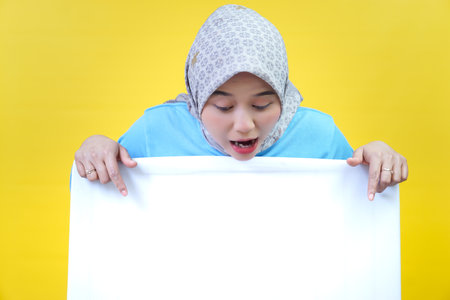 An asian woman in a hijab and blue top looks down while pointing at a blank white board on a yellow background.の写真素材