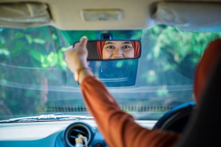 An asian muslim woman in a hijab adjusts the rearview mirror inside a car, smiling as her reflection appears.の写真素材