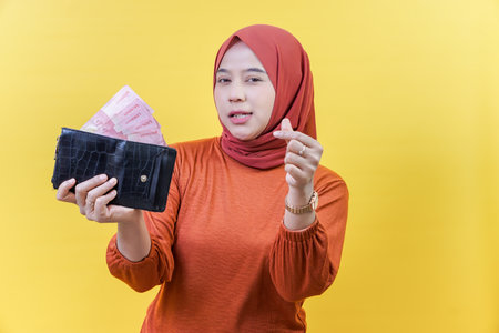 An asisan muslim woman in a hijab holds a wallet full of cash, making a hand gesture as if offering or asking for money.の写真素材