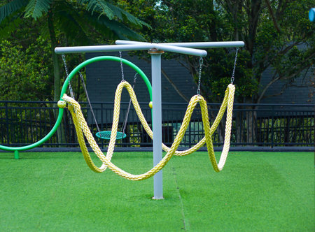 A twisted rope swing structure stands on green turf in an outdoor playground surrounded by trees.