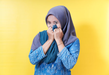 Modest woman in hijab covering face on yellow.