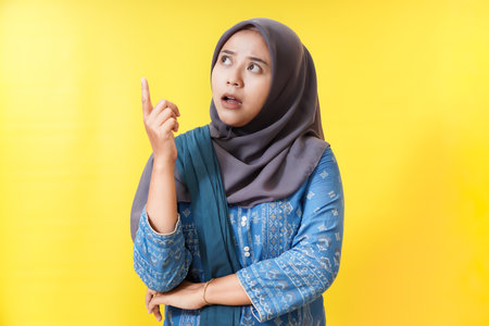 Woman in hijab pointing up with surprised expression on yellow background.の写真素材