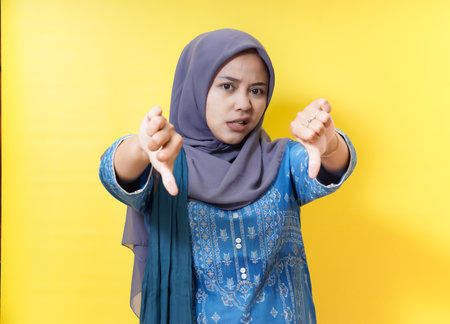 Woman in hijab gives thumbs down on yellow background.の写真素材