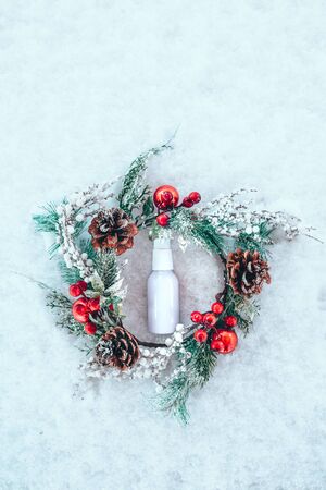 Cream serum bottle in the frame of a Christmas wreath on a background of fresh white snow. Minimal winter vacation idea. Flat lay top view composition.の写真素材