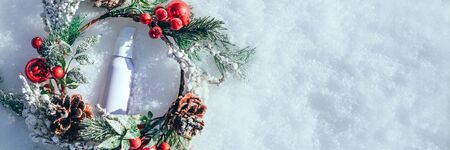 Cream serum bottle in the frame of a Christmas wreath on a background of fresh white snow. Minimal...