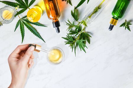 Various glass bottles with CBD oil, THC tincture and hemp leaves on a marble background. Flat lay, minimalism. Cosmetics CBD oil. Female hands holding a pipette with CBD cosmetic oilの写真素材