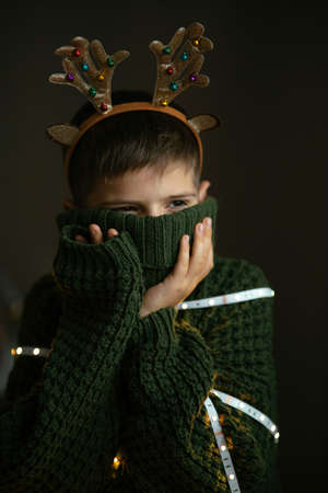 A cute boy in deer horns and a knitted Christmas sweater hides his face in the collar as in a protective mask.の写真素材