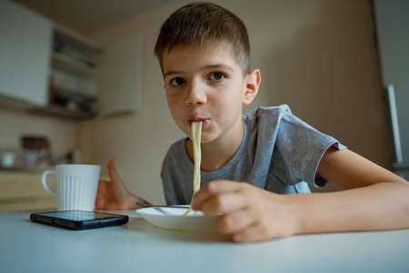 Cheerful, hungry boy eats pasta with appetite, sucks in pasta playfullyの写真素材
