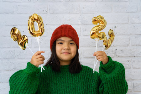 Asian child girl in ahat and green sweater close-up, in hands of golden balloons with numbers 2023