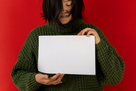 Asian woman in green sweater on red background holds paper in hands, copy spaceの写真素材