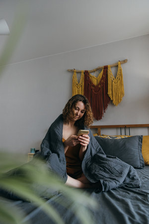 Young adult woman with smartphone sitting in bed in morning, wrapped in blanket