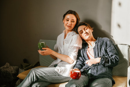 Two women spend time peacefully on the sofa on a sunny day with mugs of tea, representing friendship and relaxationの写真素材