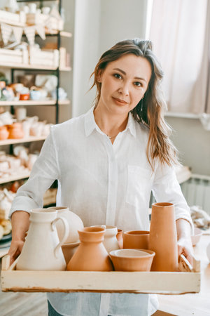 A woman master of a pottery workshop carries clay products for firing. Potter with an assortment of ceramic dishes in a bright workshopの写真素材
