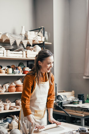 Smiling woman potter in her workshop, rolling out clay. Skilful female artisan at work, surrounded by her creations in a bright studioの写真素材