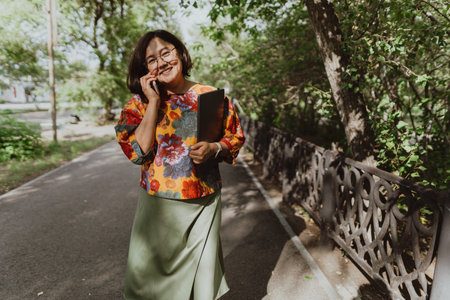 Happy woman chatting on the phone holding a laptop walking in the park Smiling freelancer wearing a colorful blouse using smartphone outdoorsの写真素材