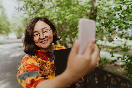 Happy woman taking a selfie with her mobile phone in a blooming park. Business casual professional taking a selfie on her cell phone while enjoying nature.の写真素材