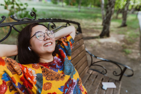 Relaxed and happy, an Asian woman enjoys her time alone on a park bench Woman in a vibrant floral shirt takes a break outdoors, smiling contentedlの写真素材