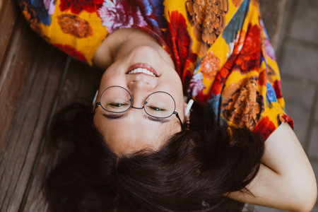 Asian woman lying on a bench a break outdoors, smiling contentedly. Joyful young woman enjoying a peaceful moment lying on a bench on a park with musicの写真素材