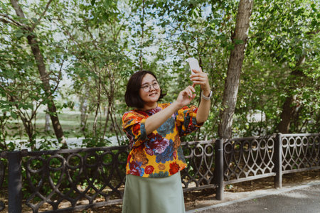 Brightly dressed woman with tech gadgets taking a selfie on sunny day in park. Happy woman taking a selfie with her mobile phone in a blooming parkの写真素材