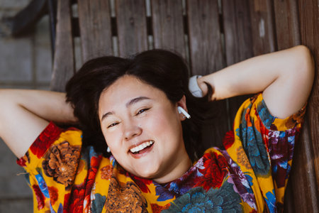 Joyful young woman enjoying a peaceful moment lying on a bench on a park with music. Taking a pause from the day, joyful woman lying on a bench in the serenity of the parkの写真素材