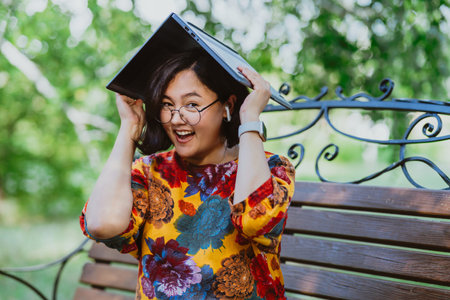 A cheerful young lady covers her head with a laptop on a sunny day. Tech-friendly environment as woman finds an unconventional use for her computerの写真素材