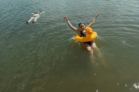 A happy child with an inflatable yellow duck is enjoying a safe swim at the lake. Her mother is...