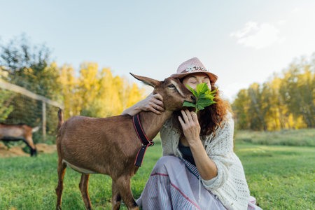 Smiling middle-aged woman in a hat bonds with the goat in peaceful rural setting. Joyful woman offers the leaves to the goat, enjoying the connection with nature on sunny dayの写真素材