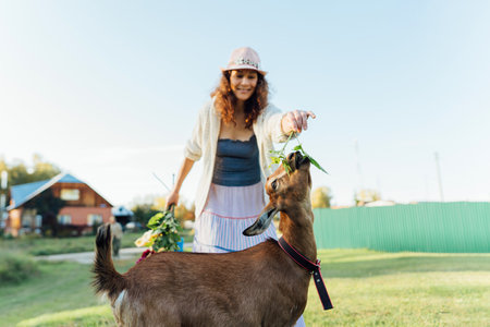 A woman is feeding a goat with fresh leaves while sitting on a farm in the countryside A content woman engages with a goat, symbolizing the peaceful and fulfilling rural lifestyle.の写真素材