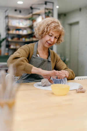 Elderly Caucasian woman sculpts clay bowl at pottery workshop, learning new skill. Senior lady enjoys working with hands, developing creativity and fine motor skillsの写真素材