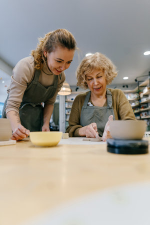 Two women work together with clay, developing skills and enjoying the artistic process. Elderly Caucasian woman learns pottery at a creative workshop with a young mentorの写真素材