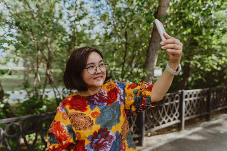 Joyful female with smartwatch capturing a selfie on sunny day in park. Young entrepreneur with digital devices capturing a selfie, staying connected in an urban green spaceの写真素材