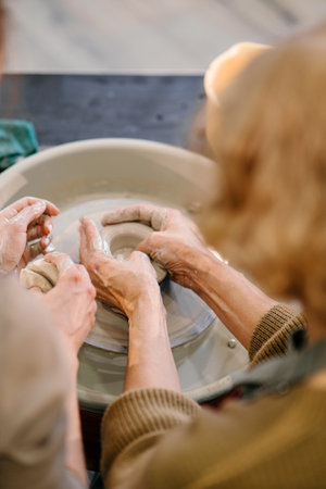 Elderly Caucasian woman practices shaping clay on a pottery wheel at a master class. A young instructor guides her hands, teaching sculpting techniques at a creative workshopの写真素材