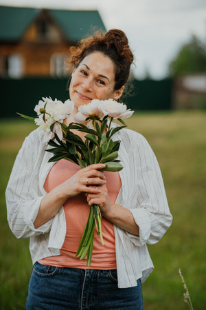 Woman standing with bouquet of blooming flowers in rustic backyard in summer. Female living simple natural life surrounded by nature and tranquilityの写真素材