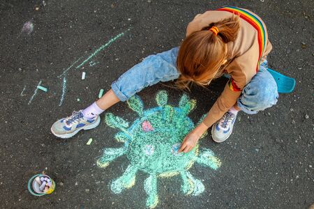 A teenage girl in a medical mask sits on a skateboard and draws a picture of the coronavirus with chalk on the pavementの写真素材