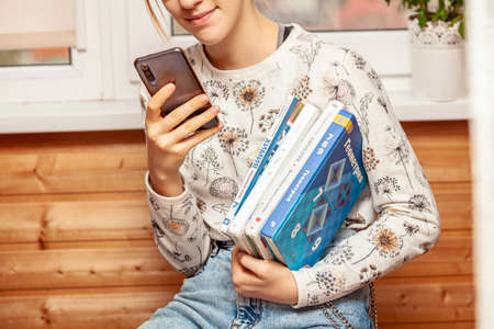 A teenage girl sits on a chair at home with textbooks and a telephone, as she is self-isolating and studying at home in Russiaのeditorial素材