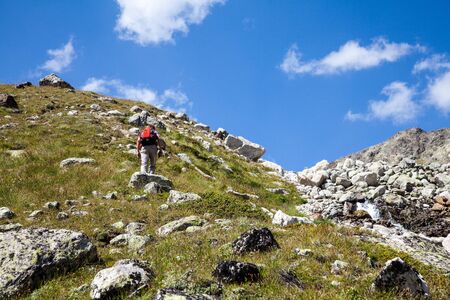 A man a tourist with a red backpack climbs up a stone slope up. A tourist carefully walks uphill. Along a mountain stream. Blue sky and white clouds. Copy space for text.の写真素材