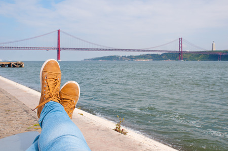 Foot an shoes. Resting near the water. Lisbon red bridge view at the background