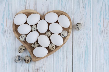 White eggs and quail eggs in wooden heart shaped plate on wooden white background, top viewの写真素材