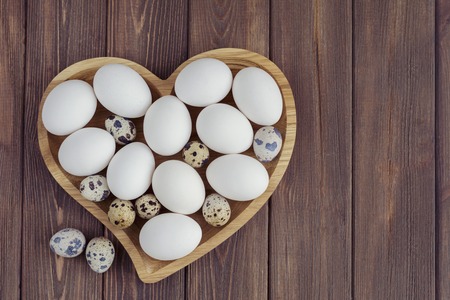 White eggs and quail eggs in wooden heart shaped plate on wooden background, easter conceptの写真素材
