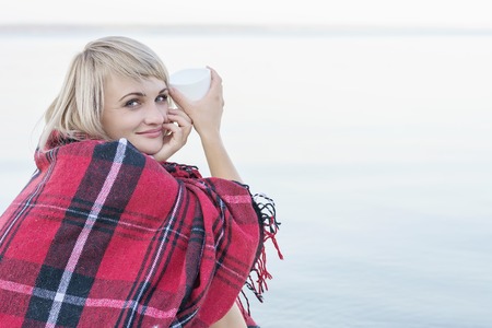 Lonely blond woman on the beach with cup of hot drink, warm red plaidの写真素材