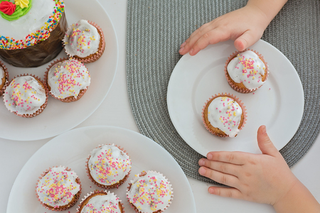 Muffins, cupcakes with confectionery on a white plate and kids hands, top view
