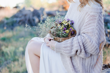 Young girl in warm light sweater is holding basket with field flowers in her hands.の写真素材