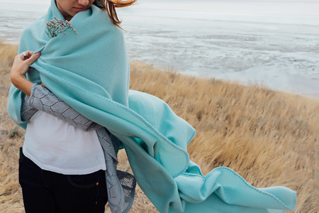 Young woman is standing on the beach in warm blanket. Windy cold weather. Cropped.