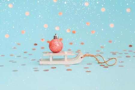 Coral glitter christmas ball on white wooden toy sleigh on blue background with confetti. Minimal styled christmas card.の写真素材