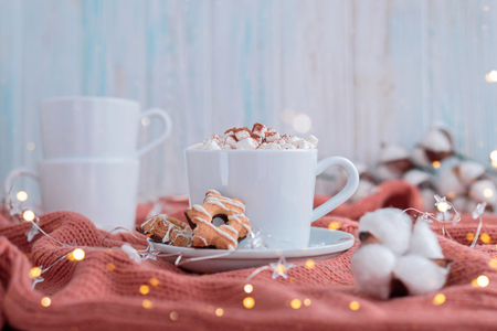 Coffee cup with marshmallow and stars cookies on warm knitted coral blanket, christmas lights and cotton flowers. Cozy and moody concept.の写真素材